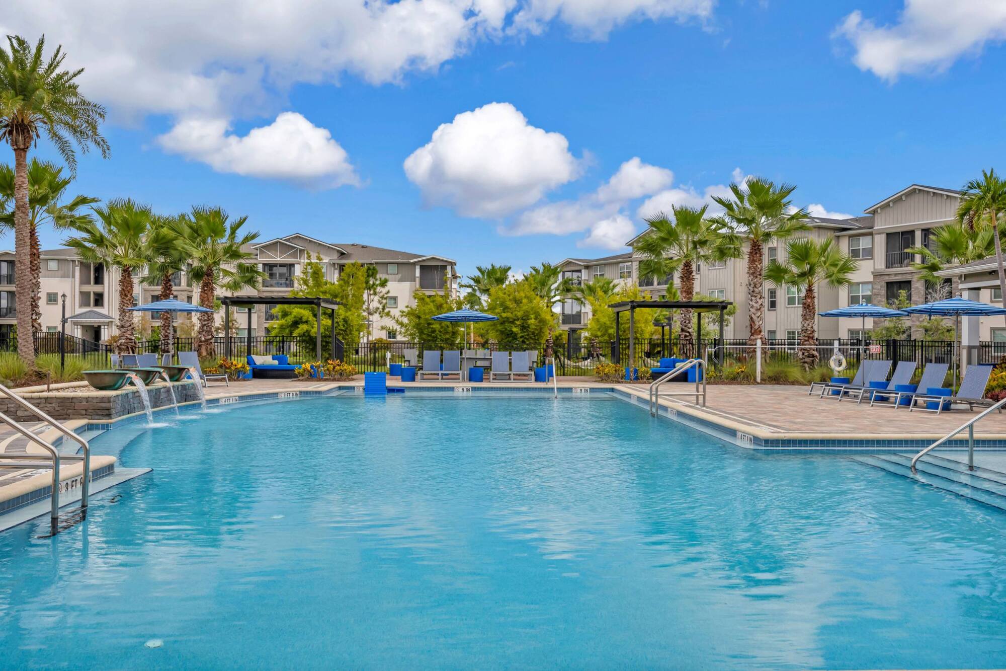 Resort-style outdoor swimming pool with lounge chairs, umbrellas, and palm trees near modern apartments.