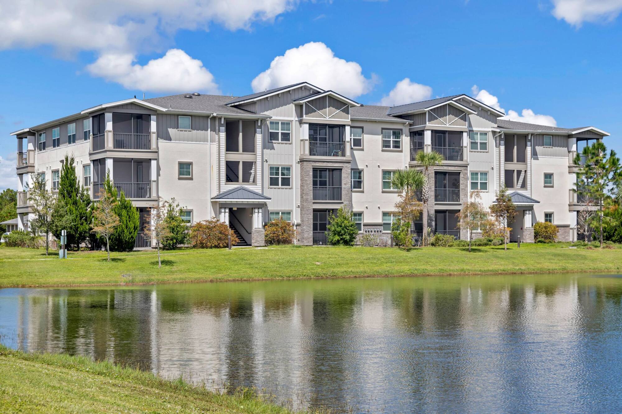 Modern three-story apartment building with balconies, reflected in a pond, under a blue sky with clouds.